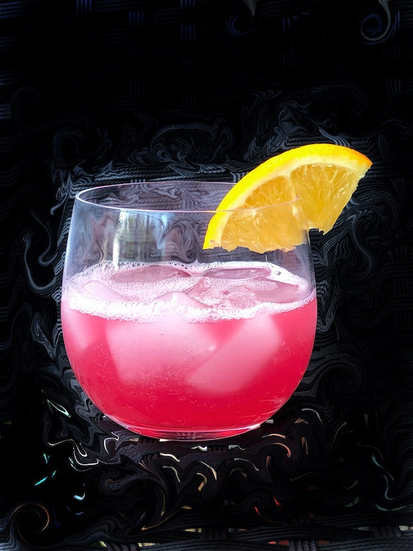Rosa Chapala Cocktail in einem runden Glas auf Eis, dekoriert mit einer Orangenscheibe vor dunklem Hintergrund.