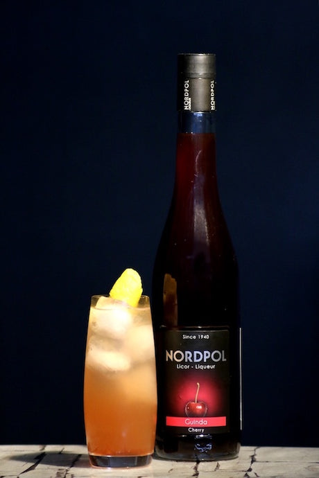 Chilcano de Guinda Cocktail im Glas mit Eis und Zitronenzeste neben Flasche Nordpol Kirschlikör vor dunklem Hintergrund