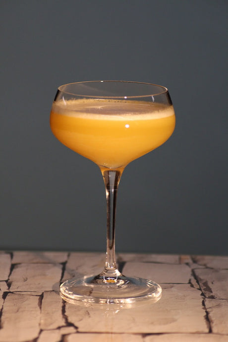 Chinese Itch Cocktail in leuchtendem Orange mit feiner Schaumkrone in einer Cocktailschale auf hellem Steinuntergrund.