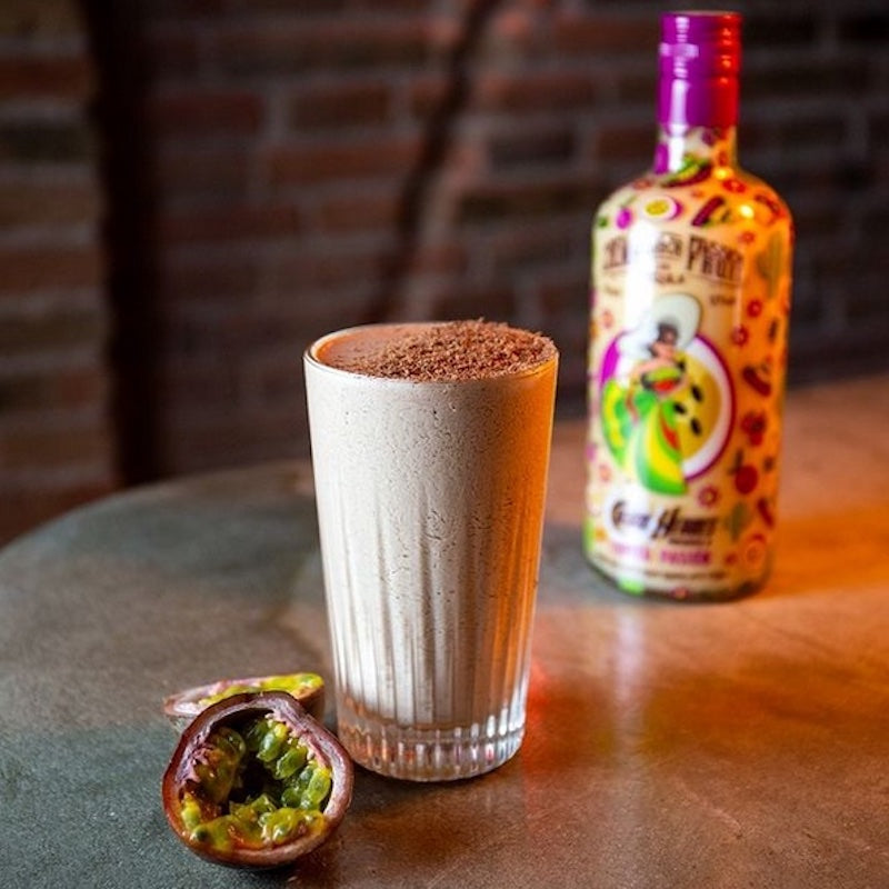 Chocopasión-Cocktail in einem hellen Glas mit Schokoraspeln, daneben aufgeschnittene Maracuja und bunte Flasche im Hintergrund.