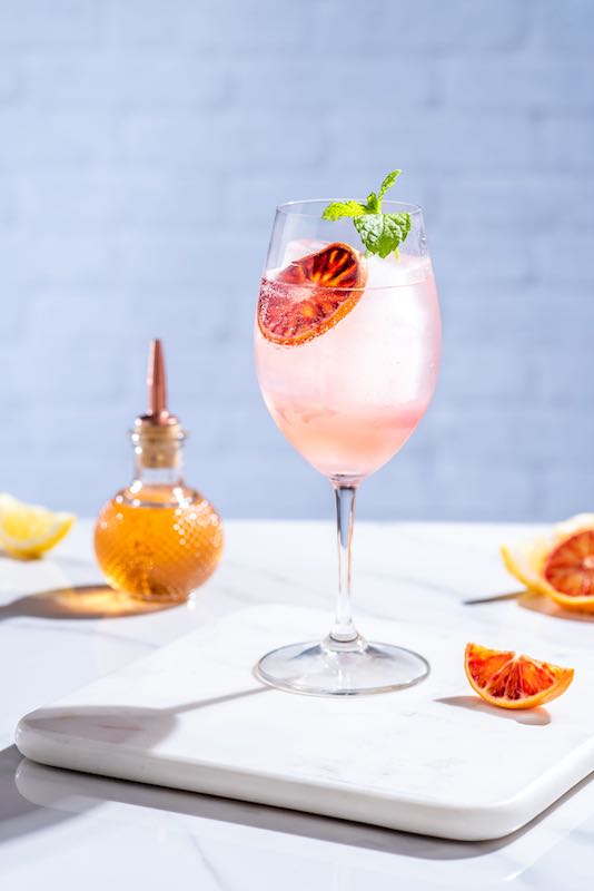 Ciao Bella Cocktail im Weinglas mit Eis, Blutorangenscheibe und Minze auf hellem Tisch