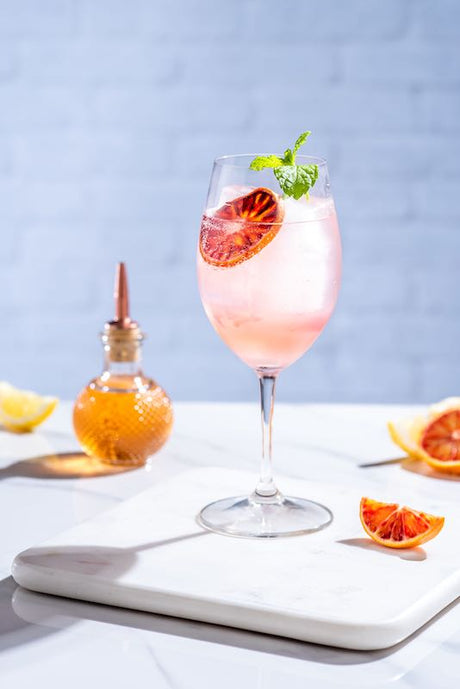 Ciao Bella Cocktail im Weinglas mit Eis, Blutorangenscheibe und Minze auf hellem Tisch