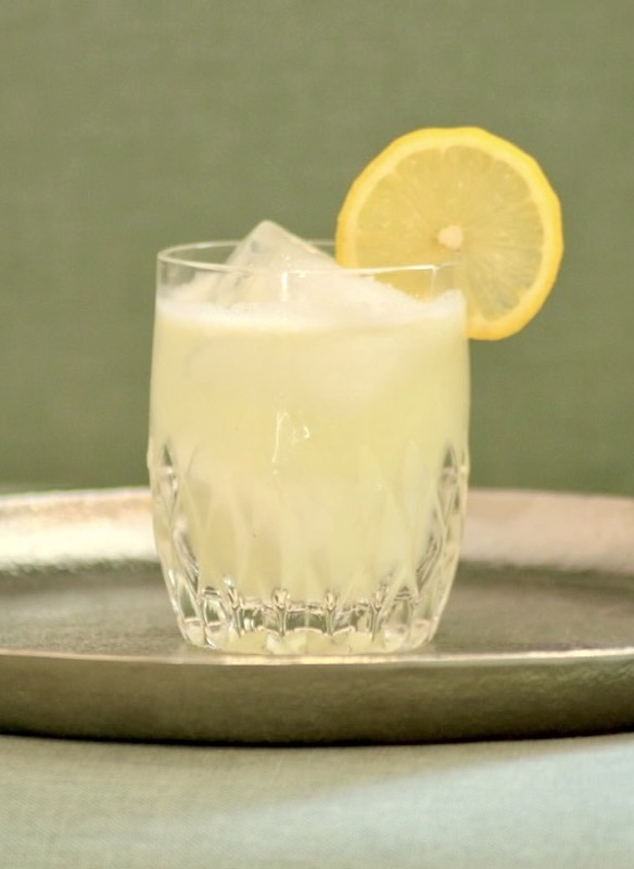 Coconut Virgin Sour Cocktail im Glas mit Eis und Zitronenscheibe auf Tablett vor grünem Hintergrund