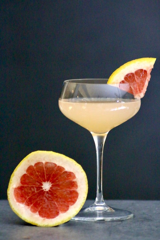 Colonial Cocktail im Glas mit Grapefruitgarnitur, daneben halbe Grapefruit auf Tisch vor dunklem Hintergrund