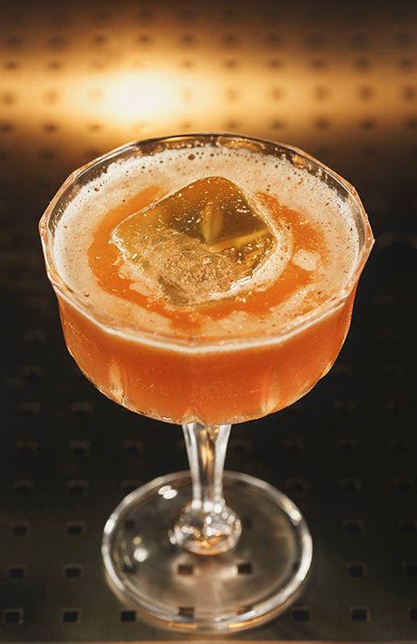 Daiquiri 5 Estrellas Cocktail in einem eleganten Glas mit großem Eiswürfel, goldbraune Farbe, vor dunklem Hintergrund.