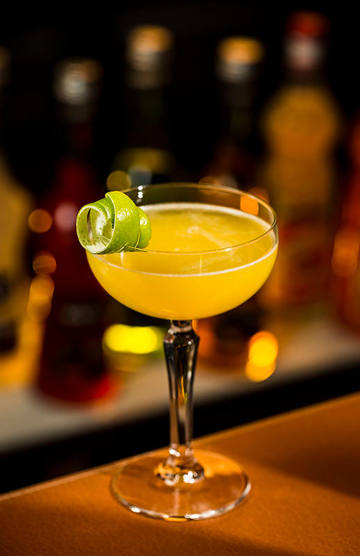 Mango Daiquiri im Coupette-Glas mit Limettengarnitur, serviert an der Bar vor stimmungsvoll beleuchtetem Hintergrund