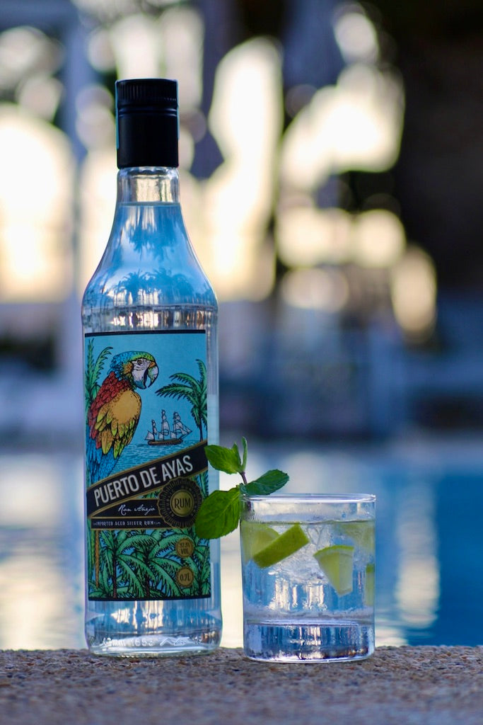 Rumflasche Puerto de Ayas neben einem Daiquiri on the Rocks mit Limettenstücken und Minze am Poolrand.