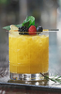 Der zerbrochene Kopf Cocktail im Old-Fashioned-Glas mit Eis, Beeren und Minze auf Tisch vor unscharfem Hintergrund
