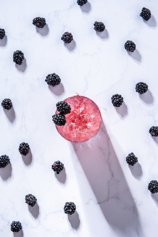 Devon Bramble Cocktail mit Crushed Ice und Brombeergarnitur, umgeben von frischen Brombeeren auf weißem Marmortisch.
