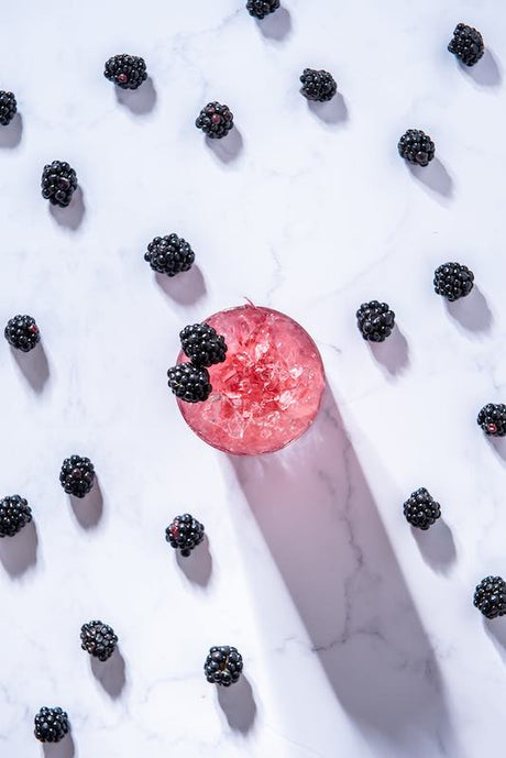 Devon Bramble Cocktail mit Crushed Ice und Brombeergarnitur, umgeben von frischen Brombeeren auf weißem Marmortisch.