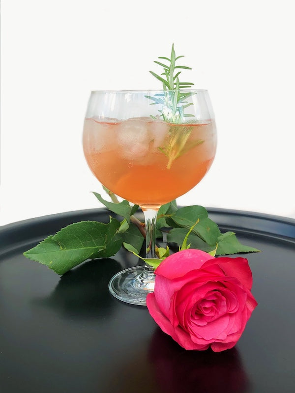 Double Rose Cocktail im Weinglas mit Eis und Rosmarin, serviert auf schwarzem Tablett mit roter Rose