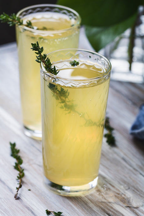 Zwei Highball-Gläser mit gelbem Fiesta d'Agrumes Cocktail und Thymianzweig als Garnitur.