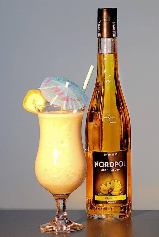 Frozen Banana Daiquiri im Hurricane-Glas mit Bananenscheibe und Schirmchen neben Flasche Bananenlikör vor hellem Hintergrund