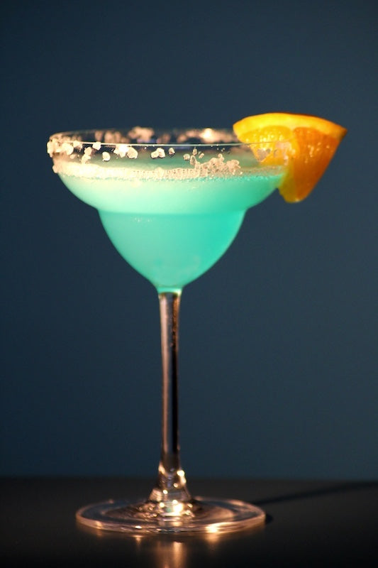 Frozen Blue Daiquiri im Cocktailglas mit Salzrand und Orangenscheibe, vor dunkelblauem Hintergrund.
