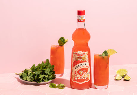 Guave-Limette-Spritz mit Sanz Guavasirop, zwei Longdrinkgläsern, Minze und Limettenspalten vor rosa Hintergrund.