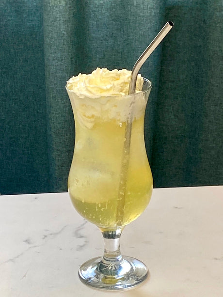 Agua de Sevilla Cocktail im Hurricane-Glas mit gelbem Drink, Schlagobershaube und Metallstrohhalm vor dunklem Hintergrund.