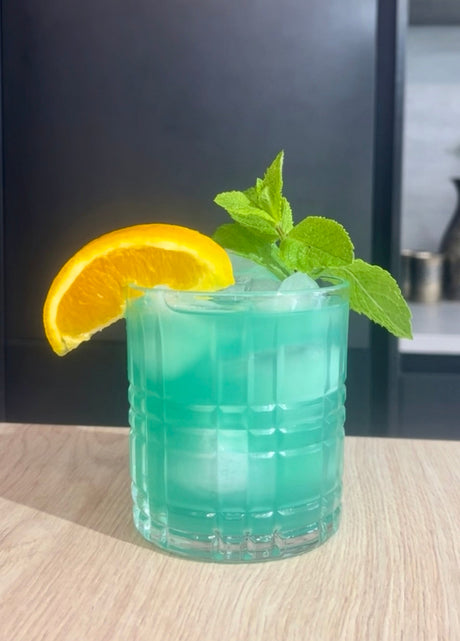 Aquamarine Cocktail auf Eis im Glas, garniert mit Orangenscheibe und frischer Minze auf Holztisch.