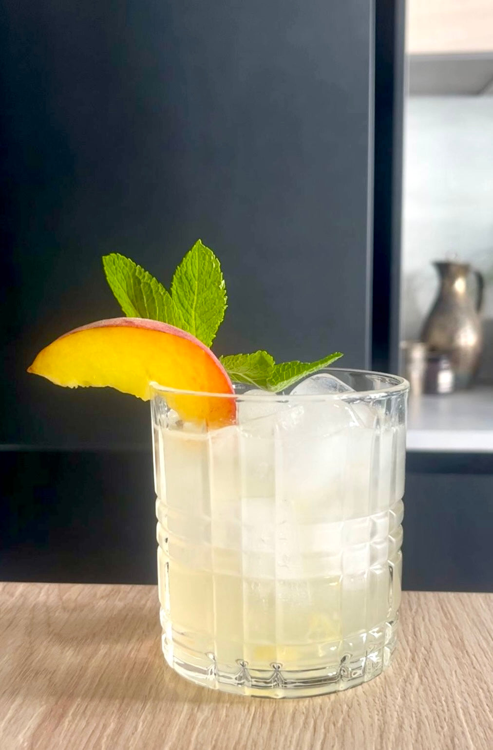 Caribbean Peach Cocktail mit Eiswürfeln, frischer Minze und Pfirsichspalte im Tumbler auf hellem Holztisch vor dunklem Hintergrund.
