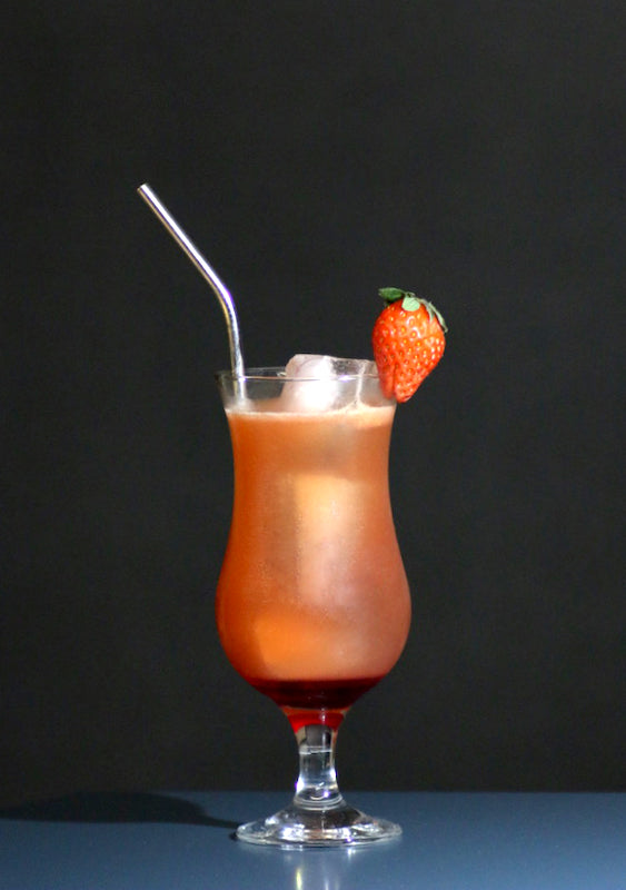 Corazonada Cocktail im Hurricane-Glas mit Eis, rötlich-oranger Farbe, Metallstrohhalm und Erdbeere als Garnitur.