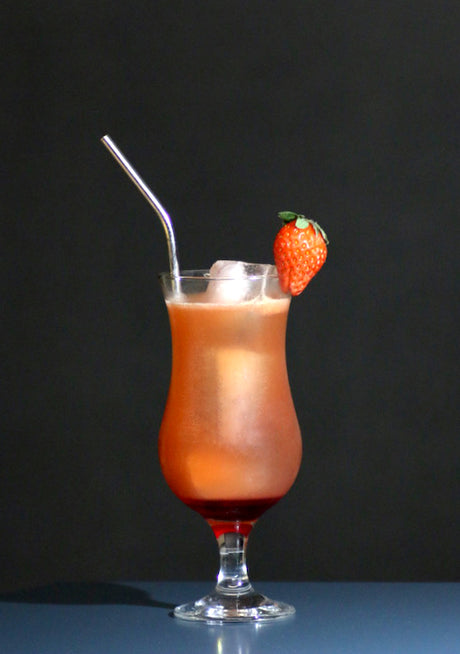 Corazonada Cocktail im Hurricane-Glas mit Eis, rötlich-oranger Farbe, Metallstrohhalm und Erdbeere als Garnitur.