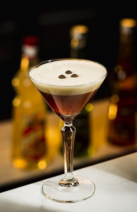 Haselnuss Espresso Martini im Martini Glas mit cremiger Schaumschicht und Kaffeebohnen auf Bar im Hintergrund