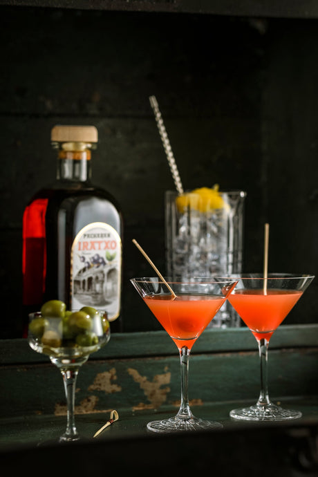 Hemingway Cocktail mit Pacharán, serviert in zwei Cocktailgläsern mit Holzstäbchen, daneben grüne Oliven und eine Flasche Pacharán.