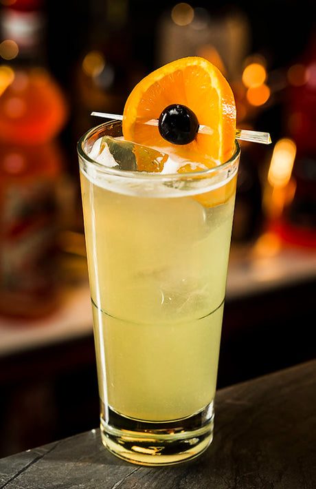 Jalisco Collins Cocktail mit Orangenscheibe und schwarzer Kirsche im Highball-Glas auf einer Bartheke.