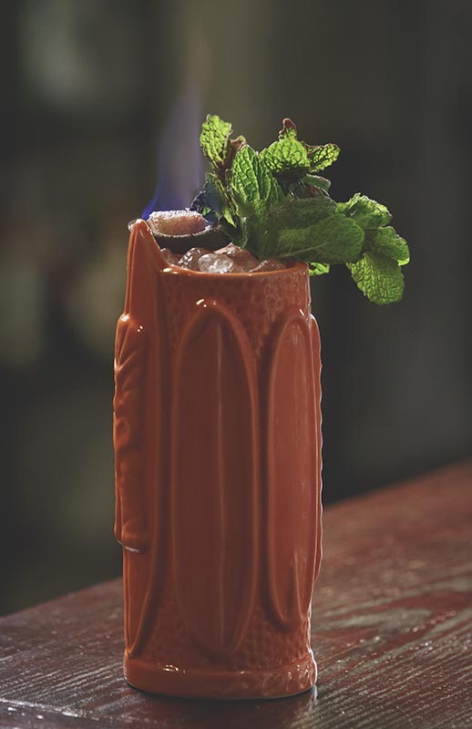 Jano Banano Tiki Cocktail in einem hohen Keramikbecher mit Crushed Ice, Minze und flambierter Garnitur.