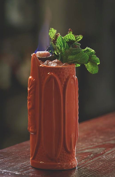 Jano Banano Tiki Cocktail in einem hohen Keramikbecher mit Crushed Ice, Minze und flambierter Garnitur.