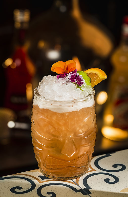 Jungle Bird Cocktail im Tiki-Glas mit Crushed Ice, Limettenscheibe, Orangenschnitz und Blüten-Deko auf einer gemusterten Barplatte.