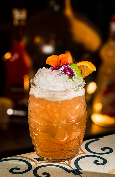Jungle Bird Cocktail im Tiki-Glas mit Crushed Ice, Limettenscheibe, Orangenschnitz und Blüten-Deko auf einer gemusterten Barplatte.
