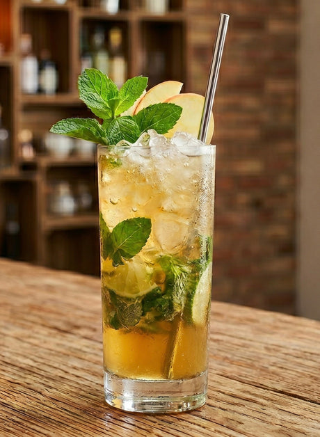 Apfel-Karamell-Mojito im Highballglas mit Crushed Ice, Minze, Limette, Apfelscheiben und Metallstrohhalm auf Holzbar