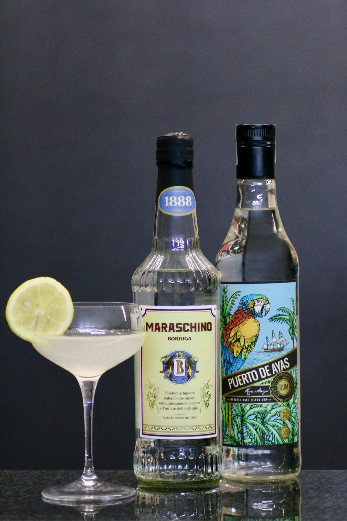 Flaschen von 'Puerto de Ayas' Rum und 'Maraschino Bordiga' Likör stehen neben einem Coupe-Glas mit King's Jubilee Cocktail.