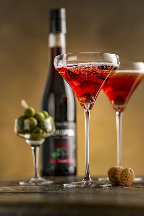 Kir Royale Cocktail mit Cassis Likör Nordpol, serviert in Sektgläsern, dazu grüne Oliven und geöffnete Champagnerflasche.
