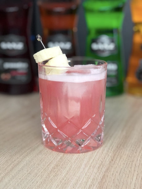 Lana Banana alkoholfreier Cocktail im Rocks Glas mit Eis und Bananenscheiben auf Tisch mit Flaschen im Hintergrund