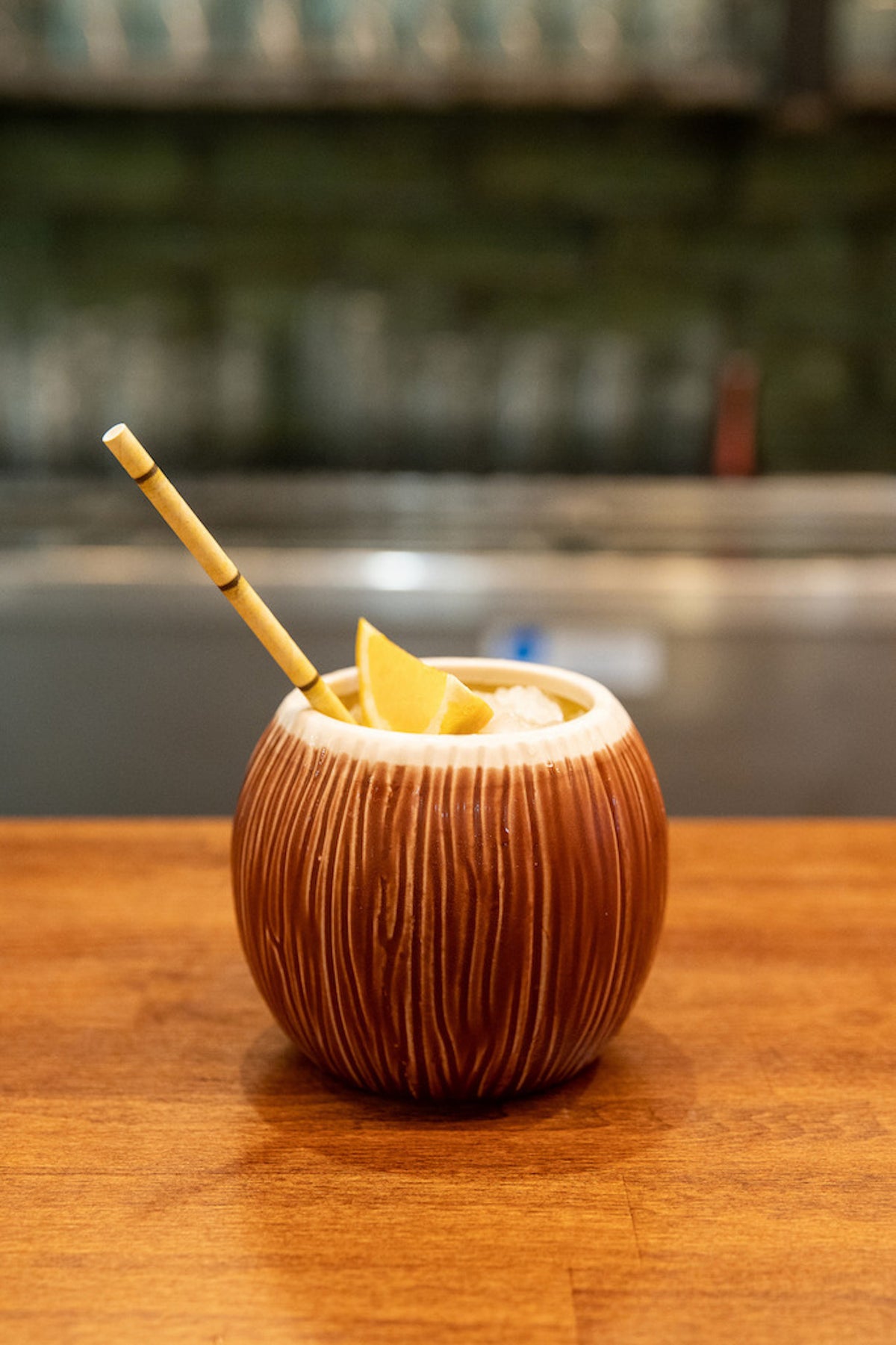 Lapu Lapu Cocktail im braunen Tiki-Glas mit Eis, Zitronenspalte und Bambusstrohhalm auf Holzbar