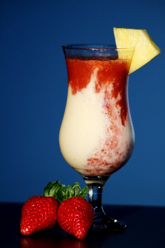 Glas Lava Flow Cocktail mit Ananas-Kokos-Creme und Erdbeerwirbel, garniert mit Ananasstück und Erdbeeren, blauer Hintergrund.