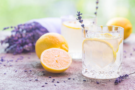 Glas mit Lavendel-Gin-Tonic, Zitronenscheibe und Lavendelzweig, daneben frische Zitronen und Lavendelblüten auf dem Tisch