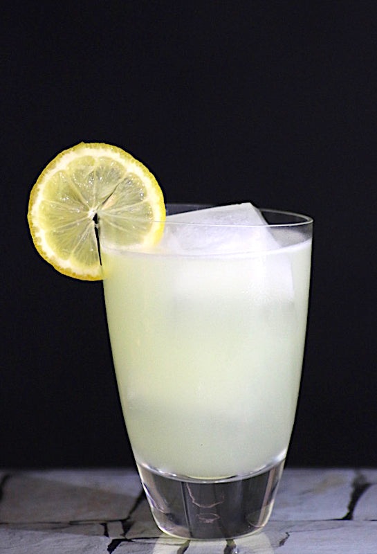 Trüber, hellgelber Lemon Burst Cocktail im Tumbler-Glas mit Eis und Zitronenscheibe vor dunklem Hintergrund.