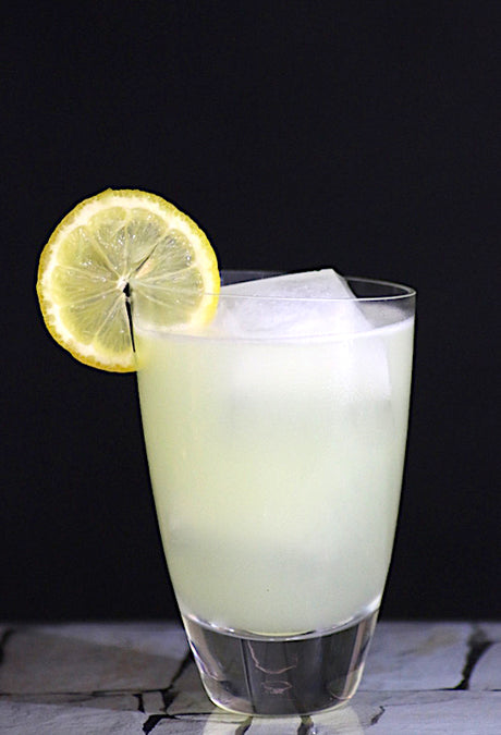Trüber, hellgelber Lemon Burst Cocktail im Tumbler-Glas mit Eis und Zitronenscheibe vor dunklem Hintergrund.