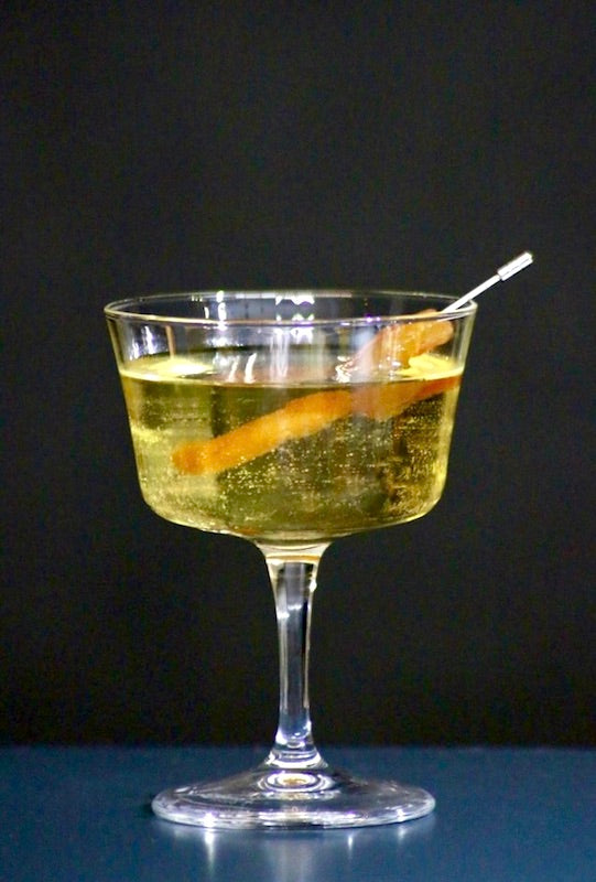 Linda Fiesta Cocktail in einem Champagnerglas, dekoriert mit einer Orangenscheibe, vor schwarzem Hintergrund.