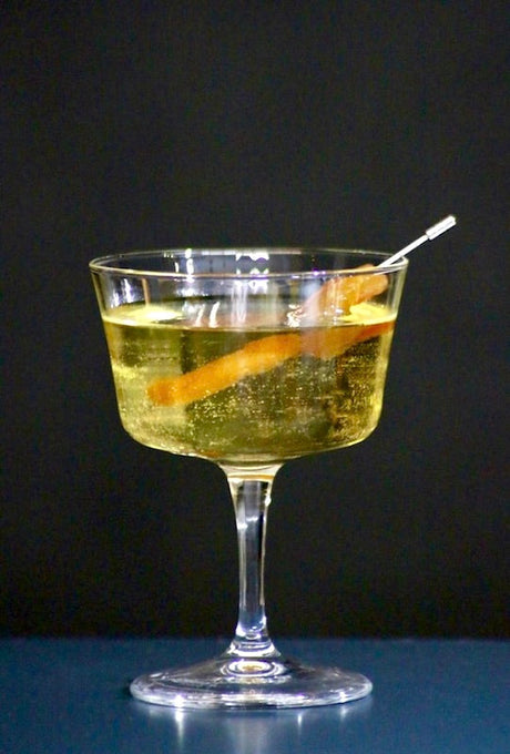 Linda Fiesta Cocktail in einem Champagnerglas, dekoriert mit einer Orangenscheibe, vor schwarzem Hintergrund.