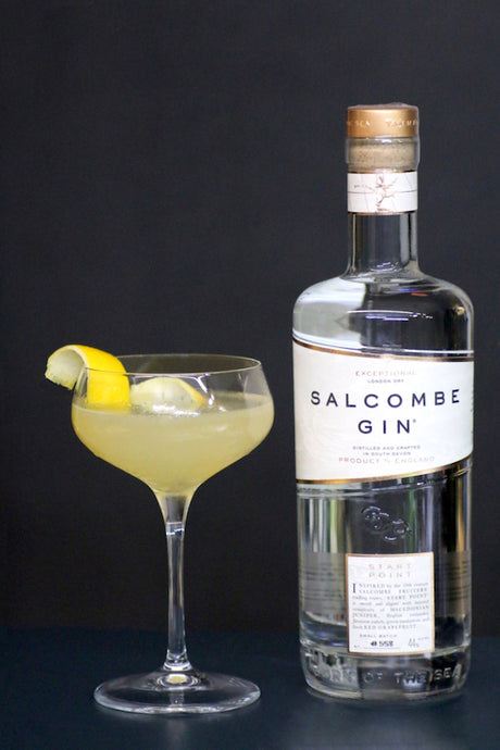 London Calling Cocktail mit Salcombe Gin in einer Coupette mit Zitronenzeste und klarer Eiskugel vor schwarzem Hintergrund.