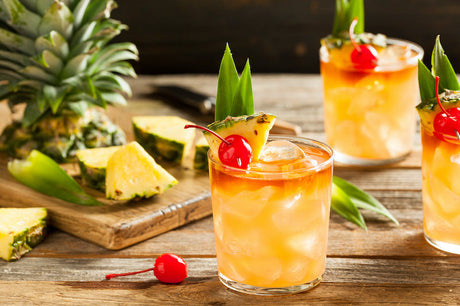 Mai Tai Cocktail, dekoriert mit Ananasstück, Kirsche und Ananasblatt, serviert auf einem Holzbrett mit frischen Ananasstücken.