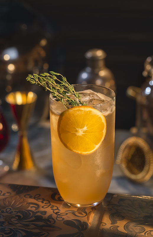 Mandarine Thymian Collins Cocktail auf Eis in einem Highball-Glas, garniert mit einer Orangenscheibe und frischem Thymian.