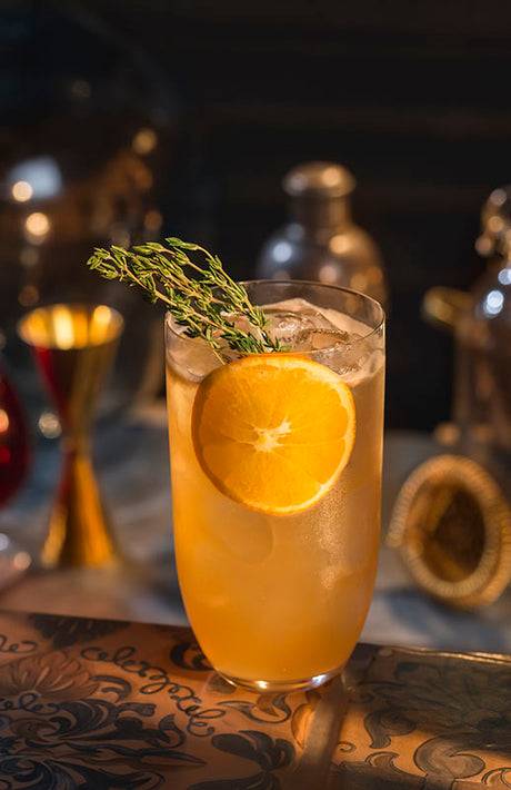 Mandarine Thymian Collins Cocktail auf Eis in einem Highball-Glas, garniert mit einer Orangenscheibe und frischem Thymian.