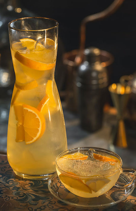Mango-Tango-Sangria in einer Karaffe und einem Glas, gefüllt mit Eiswürfeln und Orangenscheiben, vor dunklem Hintergrund.