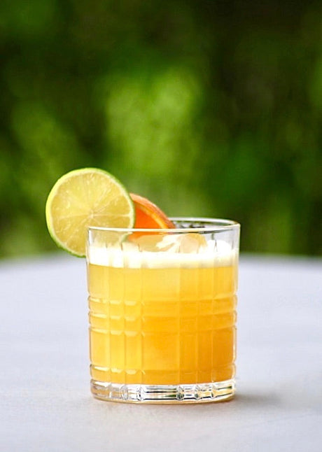 Tropical Fling Cocktail in einem Glas mit Eis, garniert mit Limetten- und Orangenscheibe, vor grünem Hintergrund.