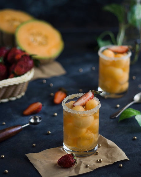Melo'politan Cocktail in zwei Gläsern mit Zuckerrand, Melonenbällchen und Erdbeere auf dunklem Untergrund