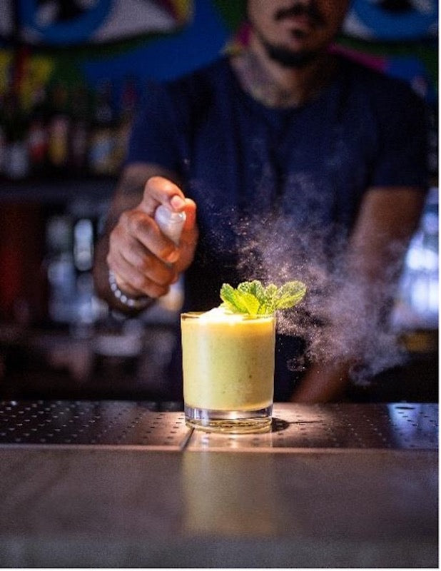 Mint & Mango Cocktail im Old Fashioned Glas, gelb, mit Minzgarnitur, Bartender im Hintergrund